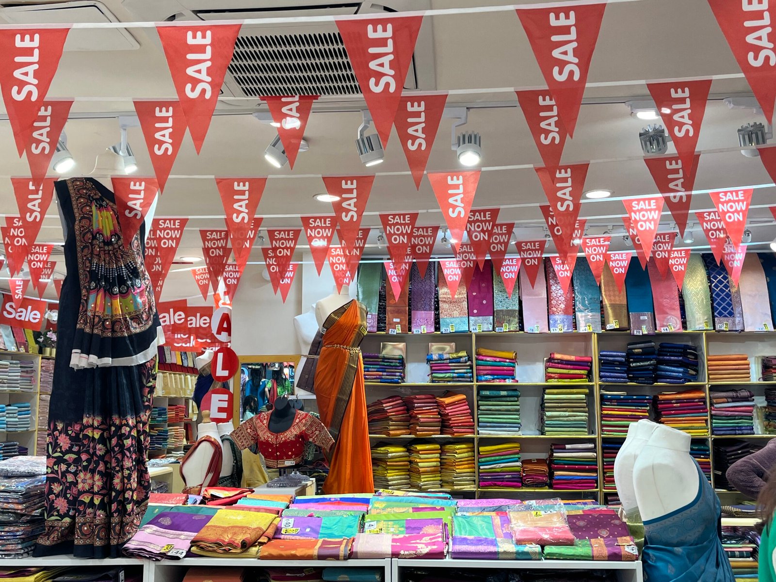 Interior of a sari shop on Ealing Road, packed with stacks of saris on shelves, featuring promotional 'Sale' banners suspended overhead
